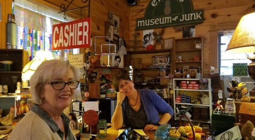 Two women smile behind a rustic counter filled with eclectic items, in a cozy shop labeled "Museum of Junk."