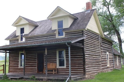 A rustic log cabin with a porch, featuring two upper windows and a sloped roof, set in a green landscape.