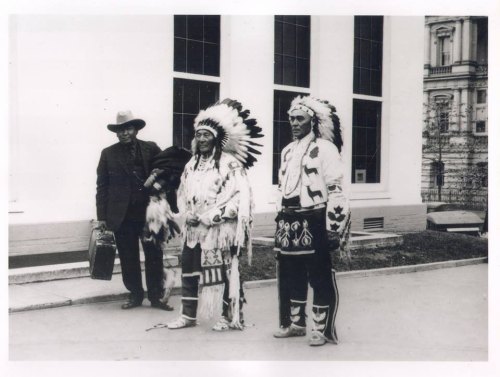 Three individuals stand outside a building; two wear traditional Native American attire, while one is in a suit.