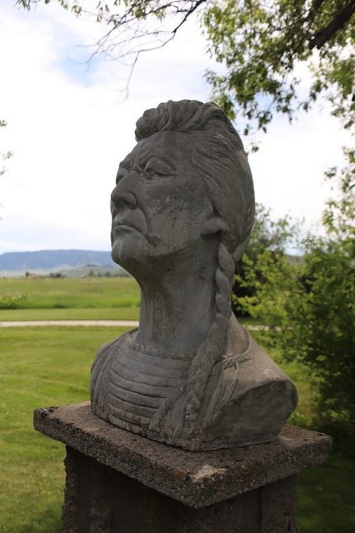 A stone bust of a person with a braid, set on a pedestal in a grassy area with trees and mountains in the background.