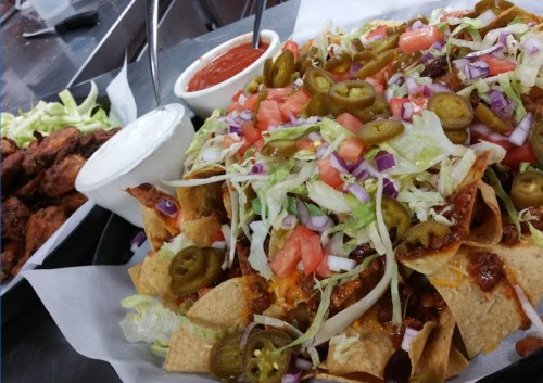 A large plate of nachos topped with cheese, lettuce, tomatoes, jalapeños, and onions, alongside a side of wings and sauces.