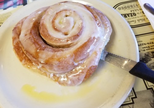 A large, gooey cinnamon roll with icing on a plate, accompanied by a knife.