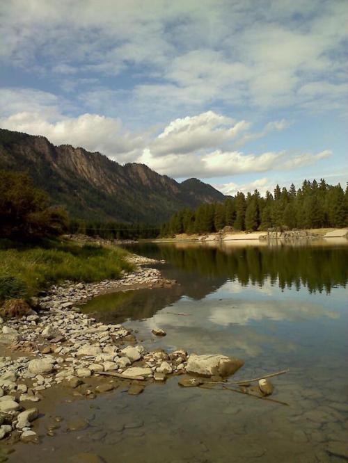 A serene river scene with rocky banks, reflecting mountains and trees under a partly cloudy sky.