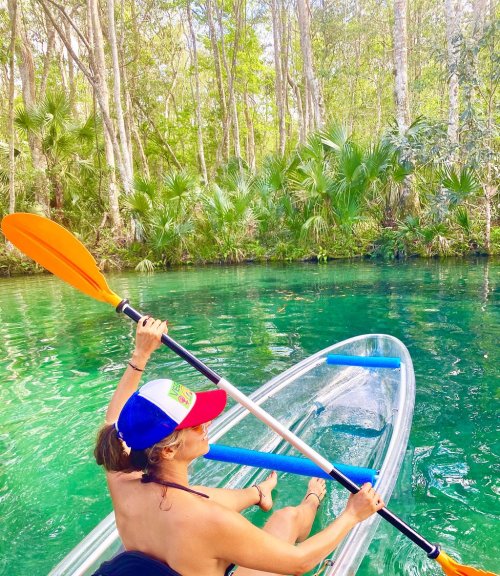 A person paddles a kayak in a lush, green river surrounded by trees and palm plants.