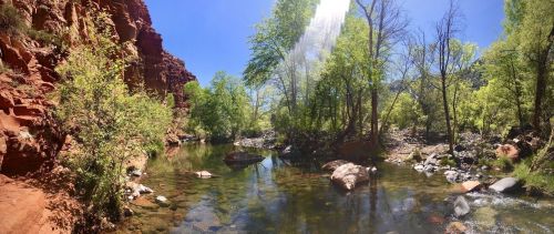 A serene river flows through a lush green landscape, surrounded by red rock formations and clear blue skies.