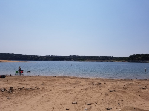 A sandy beach by a calm lake under a clear blue sky, with a person and a dog near the water's edge.