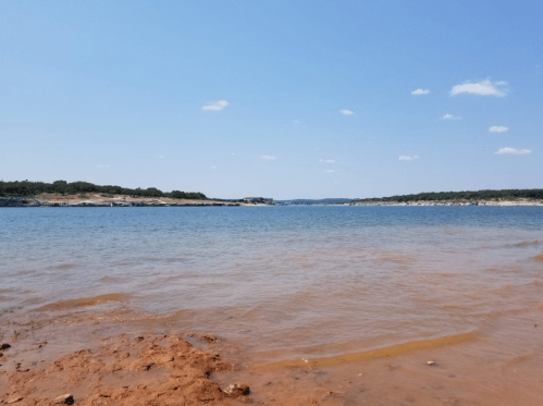 A serene lake view with clear blue water, sandy shore, and a bright blue sky dotted with a few clouds.