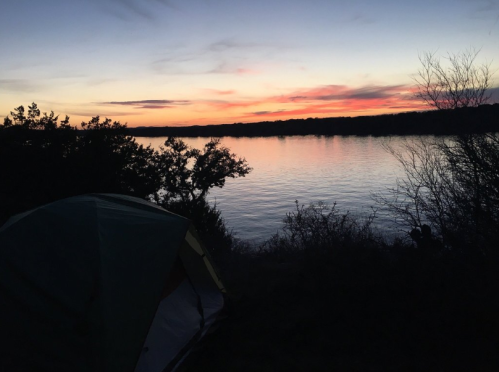 A serene lakeside view at sunset, with a tent in the foreground and colorful clouds reflecting on the water.
