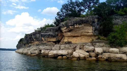 Rocky shoreline with layered cliffs and greenery under a partly cloudy sky, reflecting in calm water.