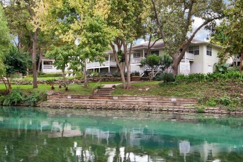 A serene riverside scene featuring green trees, a clear waterway, and a house with a porch in the background.
