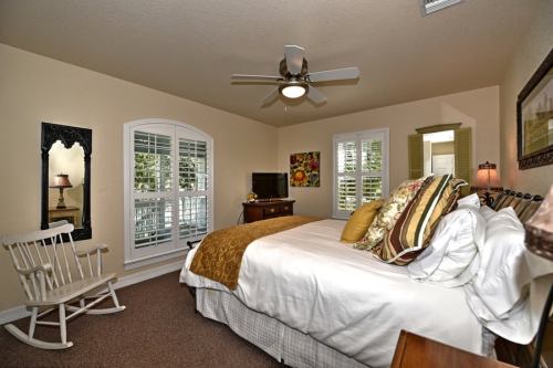 Cozy bedroom with a large bed, rocking chair, and natural light from windows, featuring warm colors and decorative accents.