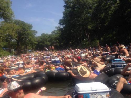 A crowded river scene with many people floating on inner tubes, surrounded by trees and a clear blue sky.