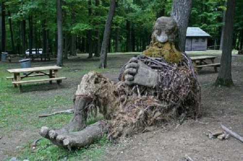 A large, moss-covered sculpture of a seated figure made from branches and natural materials in a forested area.