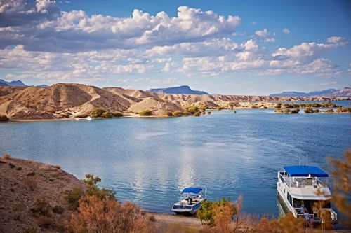 A serene lake surrounded by sandy hills and mountains, with boats docked along the shore under a blue sky with clouds.