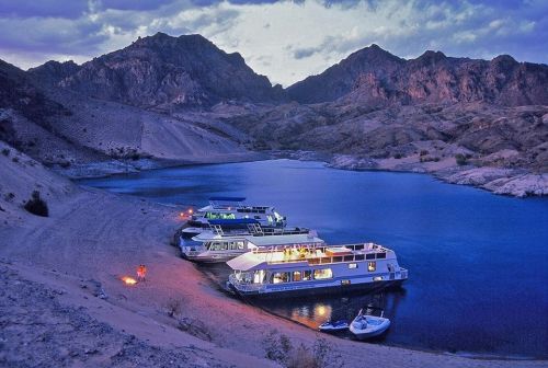 A serene lakeside scene at dusk with houseboats moored along a sandy shore, surrounded by mountains.