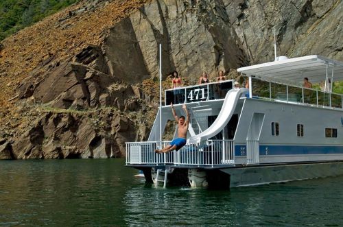 A person slides off a houseboat into the water, with friends enjoying the sun on the deck and rocky cliffs in the background.