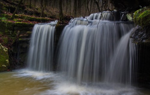 A serene waterfall cascades over rocks, surrounded by lush greenery and a calm pool below.