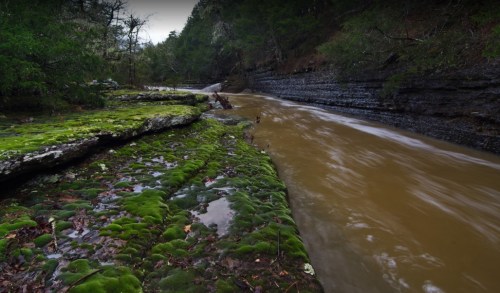 A serene river flows through a lush, moss-covered landscape, surrounded by rocky cliffs and greenery.