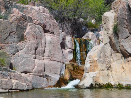 A serene waterfall cascading over pink and gray rocks, surrounded by lush greenery and clear water below.