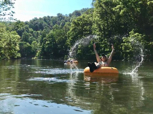 A person enjoying a summer day on a river, splashing water while floating on an orange inner tube. Lush greenery surrounds.