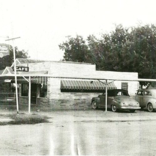 A vintage black-and-white photo of a small café with two classic cars parked outside. Trees are visible in the background.