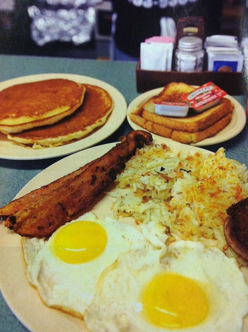 A breakfast plate with two eggs, hash browns, bacon, and toast, alongside a stack of pancakes in the background.