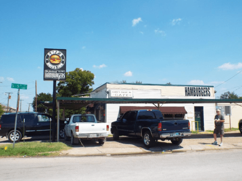 A small café with a sign reading "HAMBURGERS," featuring parked cars outside on a sunny day.