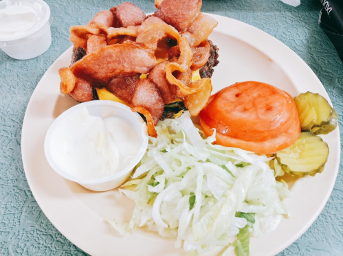 A plate with a bacon-topped burger, lettuce, tomato, pickles, and a side of ranch dressing.