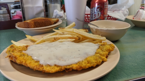 A plate with fried chicken smothered in gravy, fries, toast, and a bowl of white sauce, with a soda in the background.
