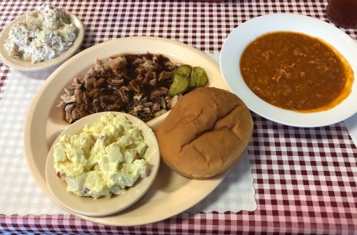A plate with pulled pork, pickles, a bun, potato salad, and a bowl of chili on a checkered tablecloth.