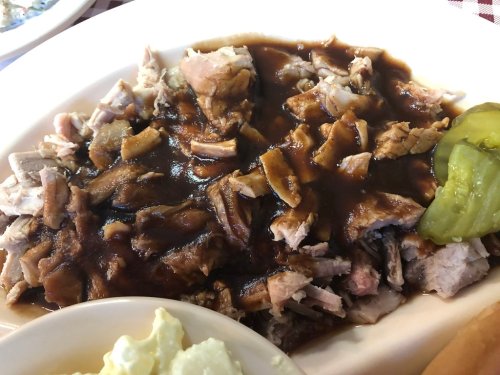 A plate of pulled pork drenched in dark sauce, garnished with pickles, alongside a side of creamy potato salad.