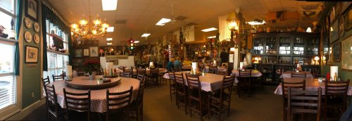A cozy restaurant interior with wooden tables, checkered tablecloths, and eclectic decor, including plates and chandeliers.