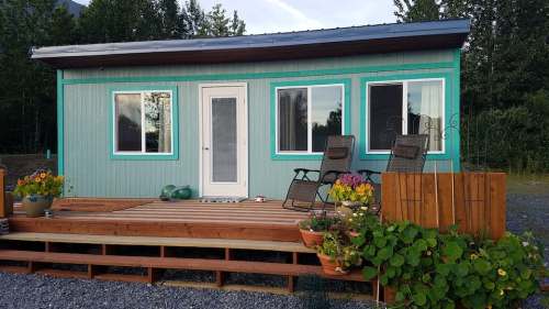 A small turquoise house with a wooden deck, potted plants, and two chairs on the porch, surrounded by greenery.