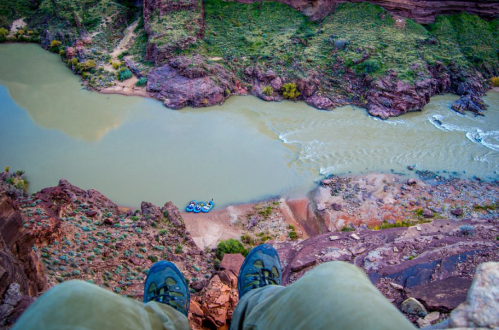 Aerial view of a river with a small raft below, seen from a rocky ledge with a person's feet in the foreground.