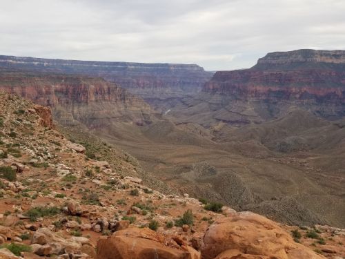 A panoramic view of the Grand Canyon, showcasing layered rock formations and rugged terrain under a cloudy sky.
