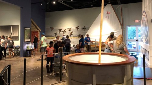 A busy museum exhibit featuring a sailboat, wildlife displays, and visitors exploring the interactive space.