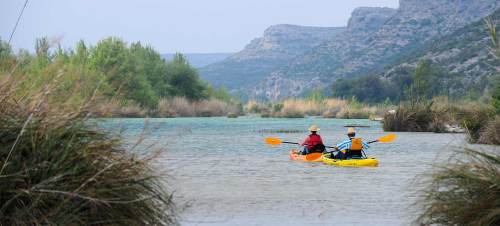 Two kayakers paddle on a calm river surrounded by lush greenery and rocky hills in the background.