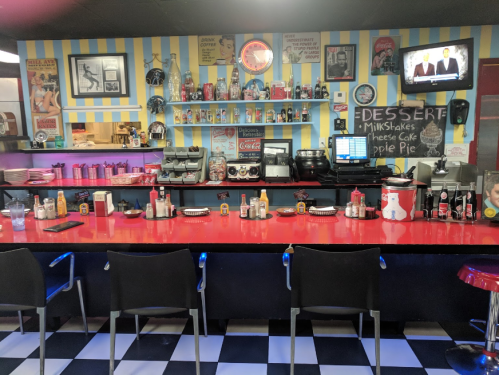 A retro diner interior with a red counter, black chairs, vintage decor, and a TV displaying news.