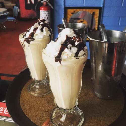 Two tall glasses of creamy milkshakes topped with whipped cream and chocolate sauce, served on a tray.