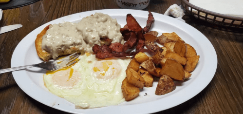 A plate of breakfast featuring eggs, biscuits with gravy, bacon, and seasoned potato wedges.