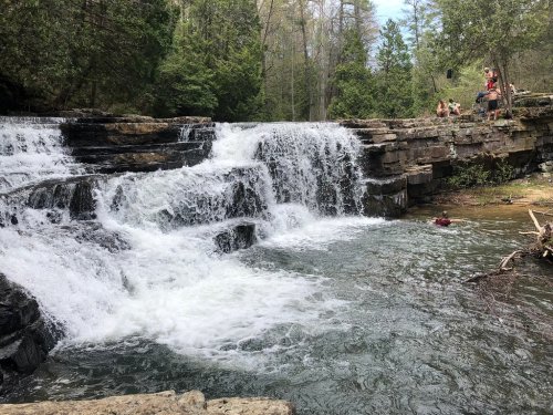 A scenic waterfall cascades over rocks into a clear pool, surrounded by lush greenery and people enjoying the outdoors.