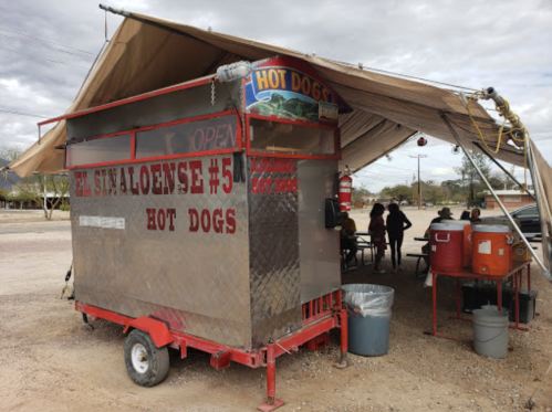 A food cart labeled "El Sinaloense #5 Hot Dogs" under a canopy, with people seated nearby.