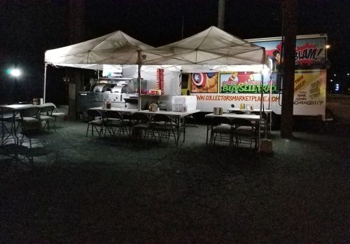 Food truck and tents set up at night, with tables and chairs in a parking lot. Brightly colored signage visible.