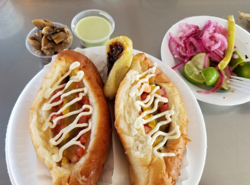 Two hot dogs topped with tomatoes and mayonnaise, served with pickled onions, lime wedges, and a green sauce on the side.