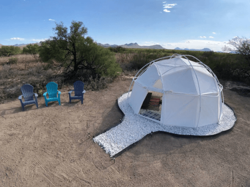 A geodesic dome tent on a sandy area, with three colorful chairs nearby and mountains in the background.