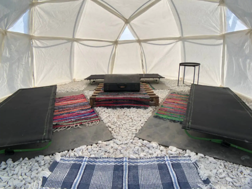 Interior of a geodesic dome with two sleeping mats, colorful rugs, and a small table on a bed of white pebbles.