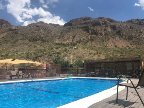 A clear blue swimming pool surrounded by mountains and a sunny sky, with lounge chairs nearby.