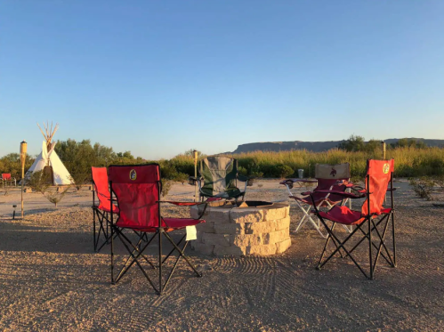 A circle of camping chairs around a stone fire pit, with a teepee and mountains in the background under a clear sky.