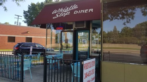 Exterior of Whistle Stop Diner with an open sign, outdoor seating, and a red awning on a sunny day.
