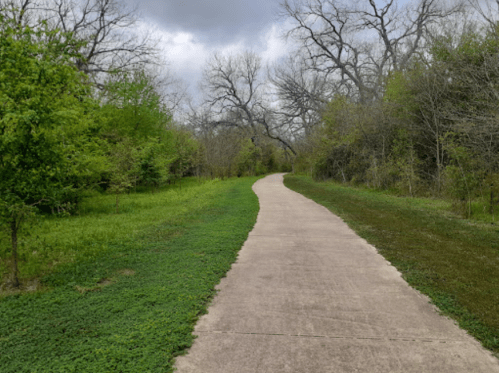 A winding concrete path through a lush green landscape with trees and cloudy skies.